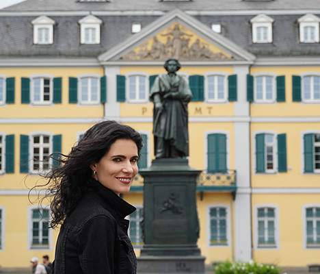 Katie Mahan stands smiling in front of a oxidized statue of a person. Behind the statue is a wide, yellow, 3-story house with green shutters.