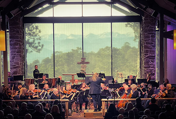 an orchestra playing in a dark auditorium. Behind them is a tall floor to ceiling window, showing a view of mountains and forests