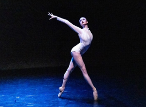 a young woman wearing a white leotard dancing on a black stage, a blue spotlight shining on her