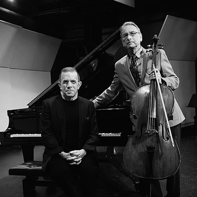 Eric Bertoluzzi, holding a cello, and Jeff Shoemaker pose in front of a piano on a stage