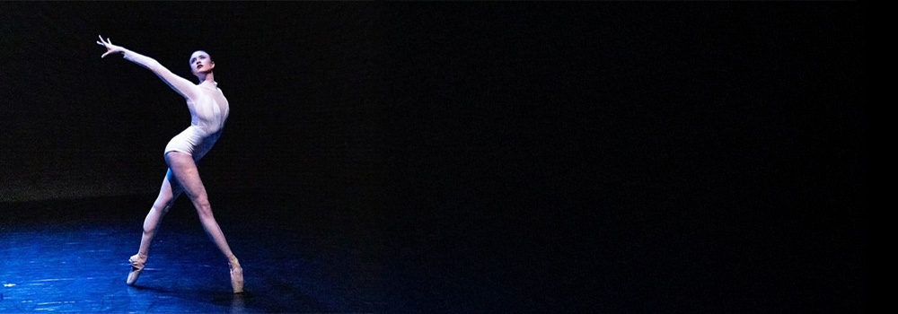 a young woman wearing a white leotard dancing on a black stage, a blue spotlight shining on her